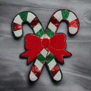May include: Two candy canes with red, white, and green sequins, crossed and tied with a red bow. The candy canes have white stripes between the red and green sections. The background is a muted gray.