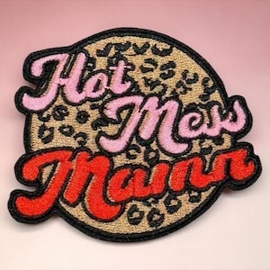 May include: Embroidered patch with the words "Hot Mess Mum" in pink and red script. The patch has a leopard print background and a black border. The patch is round.