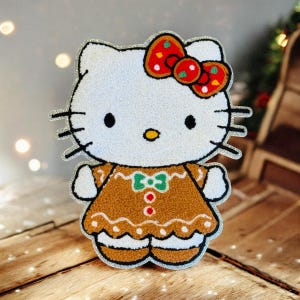 Könnte beinhalten: Ein weißer Hello Kitty-Charakter mit schwarzen Augen, Schnurrhaaren und einer gelben Nase. Sie trägt ein Kleid im Lebkuchen-Stil mit einer grünen Schleife und einer roten Schleife mit grünen Akzenten. Das Bild hat eine silberne Glitzerkontur.