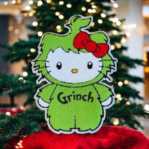 以下が含まれることがあります： 赤いリボンと黒い文字で「Grinch」と書かれた、緑と白のハローキティのデコレーションフィギュア。キラキラ光るシルバーの縁取りがあり、クリスマスツリーを背景にしています。