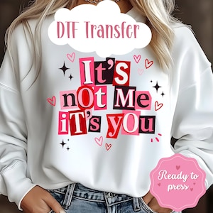 Puede incluir: Sudadera blanca con el texto "It's not Me It's You" en un estilo collage con letras rosas y rojas. El diseño incluye corazones y estrellas. También son visibles las palabras "DTF Transfer" y "Ready to press".