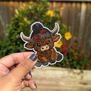 May include: An embroidered patch featuring a brown Highland cow with a fluffy texture, wearing a grey knit beanie with a pom-pom and orange pumpkin accents. The cow has curved horns, large dark eyes, and a light-colored muzzle, with a white border.