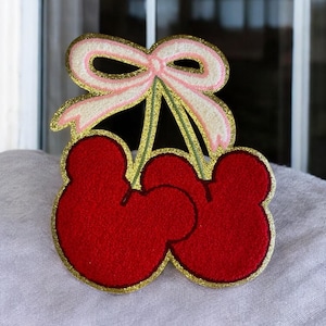 May include: A red felt cherry applique with gold glitter trim and a pink and white bow. The cherry design has a Mickey Mouse ear shape. The applique is on a white surface.