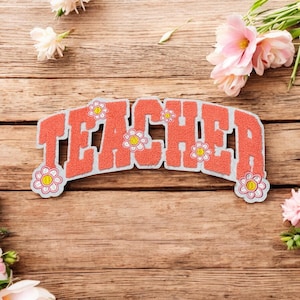 May include: A coral-colored patch with the word "TEACHER" in a bold, blocky font. The patch is outlined in white and decorated with small flower accents. The patch is on a wooden surface, with pink and white flowers.
