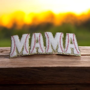 May include: A white felt applique spelling "MAMA" with red stitching and gold trim, resembling a baseball. The applique is displayed on a wooden surface with a blurred background of green and yellow.