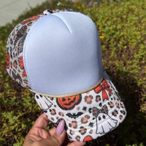 Può includere: Un cappellino trucker con pannello frontale bianco e cordoncino dorato intrecciato. La visiera e i pannelli laterali presentano un motivo ripetuto di fantasmi bianchi, zucche arancioni, pipistrelli neri, fiocchi rossi e macchie leopardate.