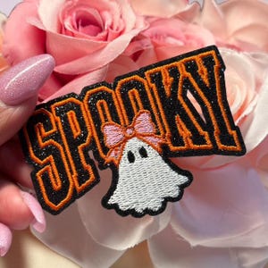 May include: An embroidered patch featuring the word SPOOKY in bold black letters with orange outlines and a glittery finish. Below the letters is a white ghost character with two black eyes, wearing a light pink bow on its head.