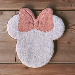 May include: A white and pink Minnie Mouse-shaped bath mat with a bow. The mat has a gold border and is made of a soft, textured material. The bow is a light pink color.
