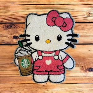 May include: A plush rug featuring the character Hello Kitty in a pink outfit, holding a Starbucks coffee. The rug has a white body, black whiskers, and a pink bow. The background is a wooden surface.