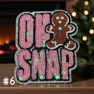 Op de afbeelding: Een decoratieve patch met de tekst OK SNAP in roze lovertjes en een bruine peperkoekfiguur. De patch heeft een zilveren glitterrand, een zwarte stoffen achterkant en kleine groene en rode knoopdetails op de peperkoekfiguur.