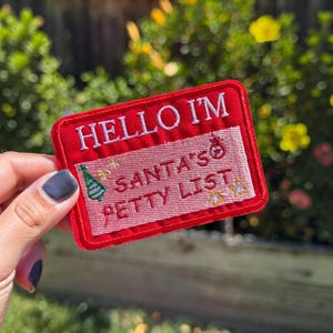 May include: A rectangular embroidered patch with a red border and a pink center. The text HELLO I'M is in white at the top, and SANTA'S PETTY LIST is in red in the center. The patch features a green Christmas tree and small yellow star icons.