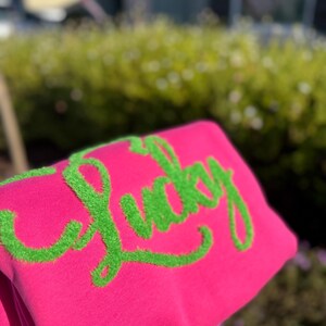 May include: A vibrant pink sweatshirt featuring the word "Lucky" embroidered in a fluffy, lime green font. The sweatshirt is folded, displaying the text. The background is blurred, with green foliage.