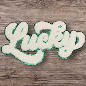 May include: White, fluffy "Lucky" sign with a green glitter outline. The word is in a cursive font, with a textured, plush appearance. The sign is set against a wooden background.