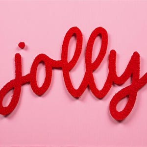 May include: Red, glittery "jolly" sign on a pink background. The word is in a cursive font, with the letters connected. The sign is likely a decorative item, possibly for the holidays.