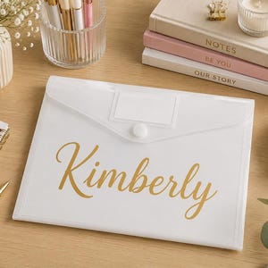 Peut inclure: Une pochette à documents en plastique blanc translucide avec une fermeture à pression et une étiquette rectangulaire vierge. Le prénom Kimberly est imprimé en lettres cursives dorées au centre de la pochette rectangulaire et plate.