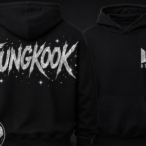 Może przedstawiać: Czarna bluza z kapturem, na której plecach widnieje napis JUNGKOOK wykonany dużą, srebrną, brokatową czcionką imitującą pociągnięcia pędzla, ozdobioną małymi gwiazdkami. Z przodu znajduje się małe, srebrne, brokatowe logo BTS na piersi.
