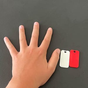 May include: Three small, rectangular tags in white and red, with a rounded top and a hole. The tags are next to a hand on a dark gray surface. The tags are likely for pet identification or keychains.