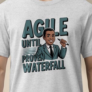 May include: Light gray t-shirt with a graphic of a smiling person holding a pen, and the words "AGILE UNTIL PROVEN WATERFALL" in teal and black. The shirt is being worn by a person.