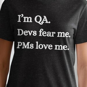 May include: Dark gray t-shirt with white text that reads "I'm QA. Devs fear me. PMs love me." The shirt is a crew neck style and is made of a soft material. The person wearing the shirt is standing in front of a white curtain and a green plant.