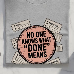 May include: A light gray t-shirt with the text "NO ONE KNOWS WHAT 'DONE' MEANS" in a pink circle. The shirt is surrounded by small cards with check marks and text. A laptop, earbuds, and a coffee mug are also visible.