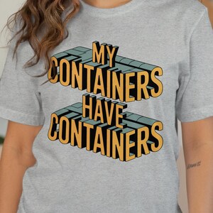 May include: A light gray t-shirt with the text "MY CONTAINERS HAVE CONTAINERS" in a 3D design. The text is in yellow and teal, with black outlines. The shirt is short-sleeved and made of a soft material.