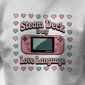 May include: White sweatshirt featuring a pink retro handheld gaming device graphic with the text "Steam Deck is my Love Language" surrounded by pink and blue hearts. The sweatshirt is a casual style.
