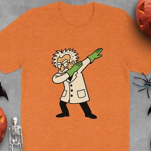 May include: Orange t-shirt featuring a cartoon scientist dabbing, wearing a white lab coat and green gloves. Halloween decorations, including pumpkins, spiders, and bats, surround the shirt, creating a festive theme.