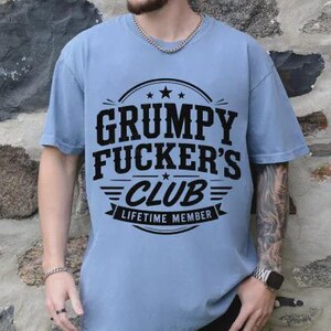 May include: A light blue t-shirt with the text "GRUMPY FUCKER'S CLUB LIFETIME MEMBER" in a black, bold font. The text is centered on the shirt, with a circular design above and a banner below. The shirt has short sleeves.