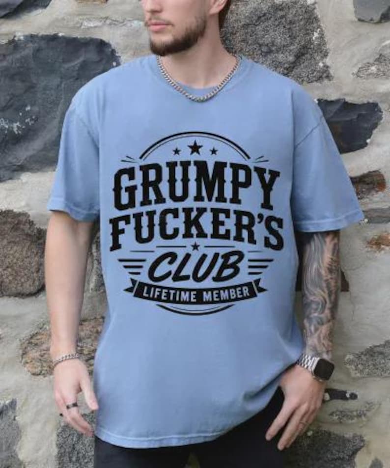 May include: A light blue t-shirt with the text "GRUMPY FUCKER'S CLUB LIFETIME MEMBER" in a black, bold font. The text is centered on the shirt, with a circular design above and a banner below. The shirt has short sleeves.