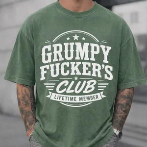 May include: An olive green t-shirt with the text "GRUMPY FUCKER'S CLUB LIFETIME MEMBER" in white. The shirt has a crew neck and short sleeves. The graphic is centered on the chest.