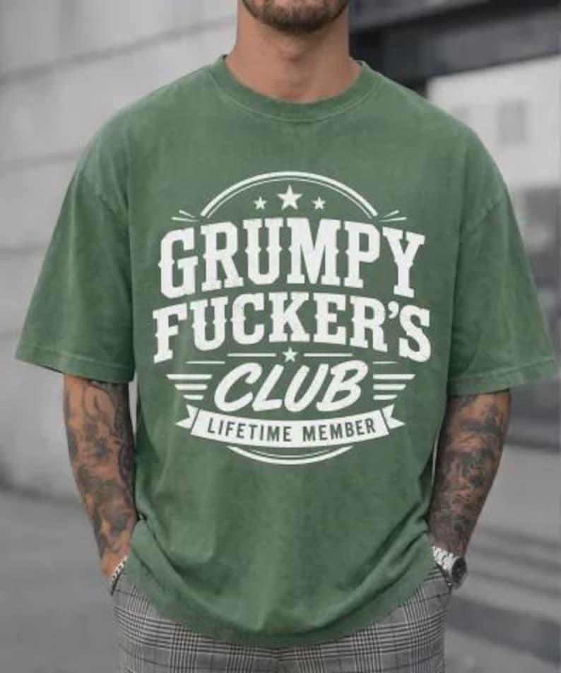 May include: An olive green t-shirt with the text "GRUMPY FUCKER'S CLUB LIFETIME MEMBER" in white. The shirt has a crew neck and short sleeves. The graphic is centered on the chest.