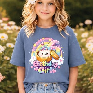 Pu&ograve; includere: Una t-shirt a maniche corte color blu polvere con la grafica di un raviolo sorridente in un cestello di bamb&ugrave;, un arcobaleno, stelle e il numero 6. Il testo "Birthday Girl" &egrave; stampato con un carattere sfumato colorato sotto il disegno.