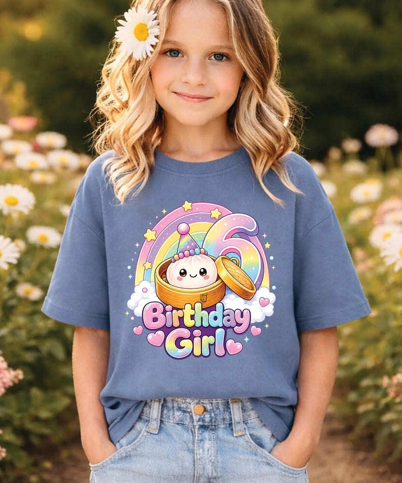 Pu&ograve; includere: Una t-shirt a maniche corte color blu polvere con la grafica di un raviolo sorridente in un cestello di bamb&ugrave;, un arcobaleno, stelle e il numero 6. Il testo "Birthday Girl" &egrave; stampato con un carattere sfumato colorato sotto il disegno.