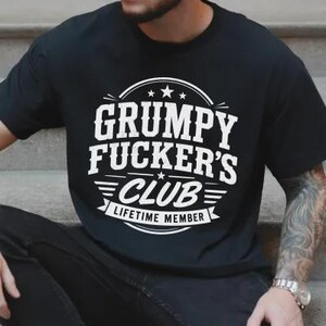 May include: Black t-shirt with the text "GRUMPY FUCKER'S CLUB LIFETIME MEMBER" in white, featuring a circular design with stars and stripes. The shirt is a casual wear item.