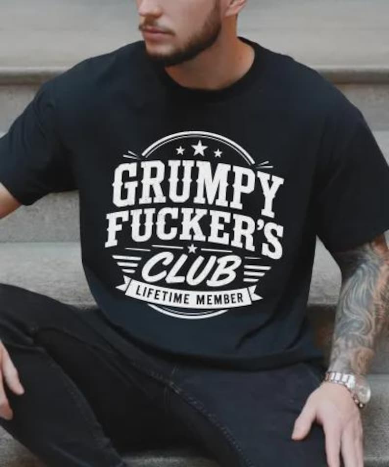 May include: Black t-shirt with the text "GRUMPY FUCKER'S CLUB LIFETIME MEMBER" in white, featuring a circular design with stars and stripes. The shirt is a casual wear item.