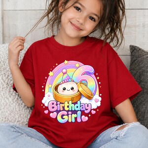 Pu&ograve; includere: Una t-shirt rossa a maniche corte con la grafica di un raviolo sorridente in un cestello per la cottura a vapore in bamb&ugrave;, un grande numero 6, un arcobaleno e la scritta "Birthday Girl" in lettere colorate, circondata da stelle e cuori rosa.