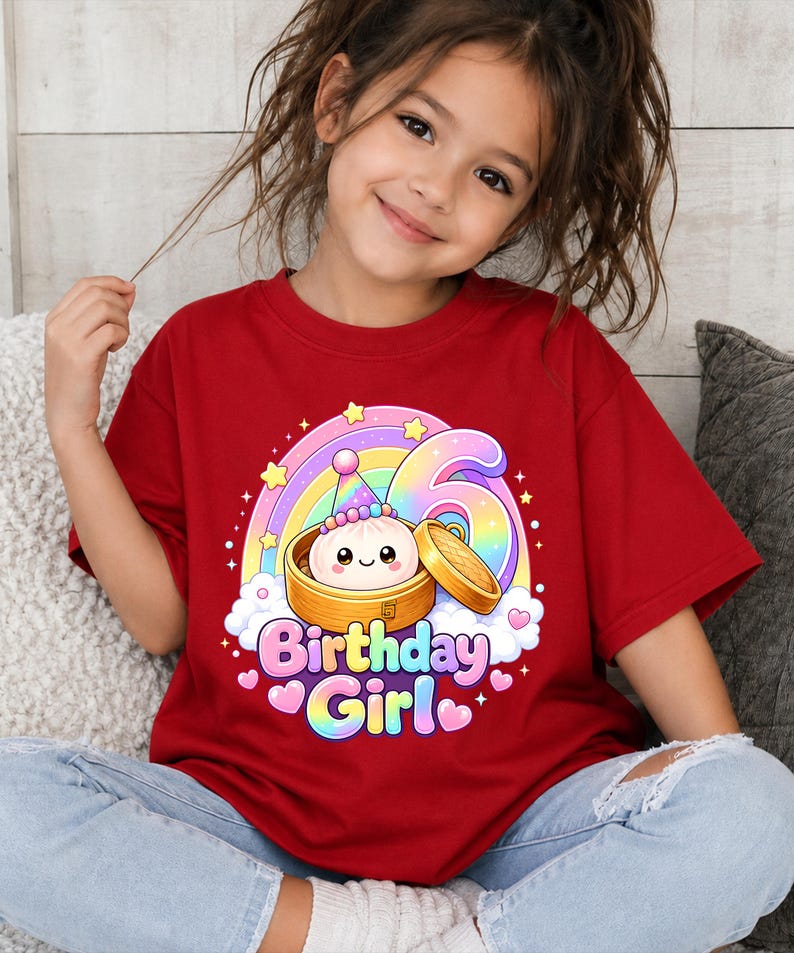 Pu&ograve; includere: Una t-shirt rossa a maniche corte con la grafica di un raviolo sorridente in un cestello per la cottura a vapore in bamb&ugrave;, un grande numero 6, un arcobaleno e la scritta "Birthday Girl" in lettere colorate, circondata da stelle e cuori rosa.
