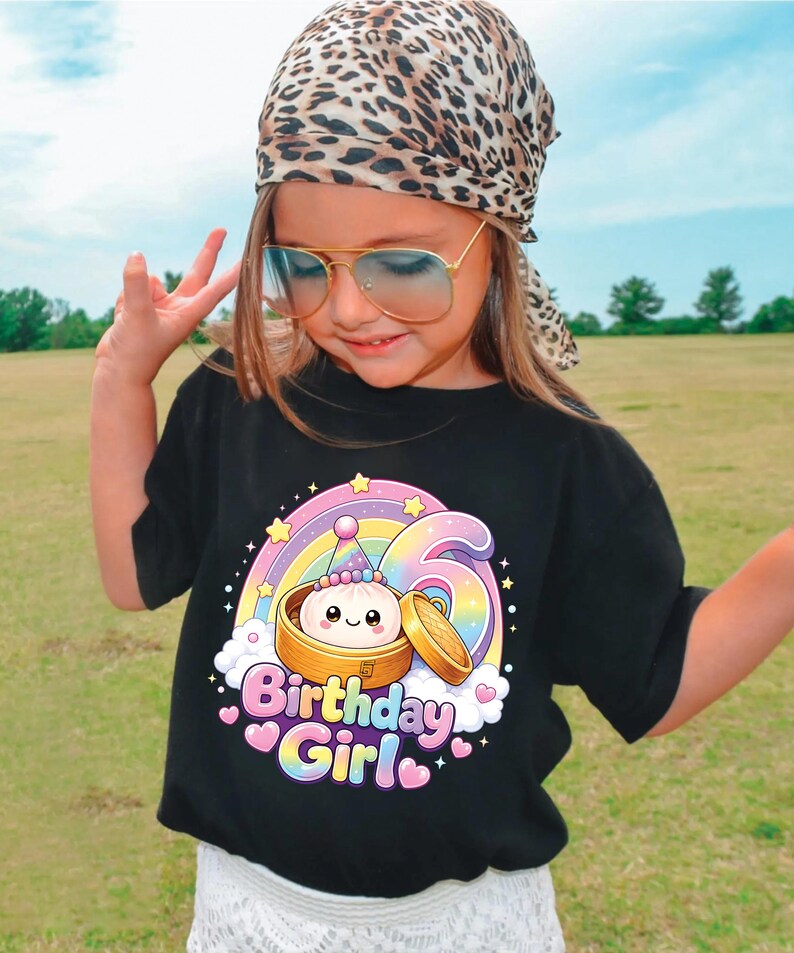 Pu&ograve; includere: Una t-shirt nera con la grafica di un grazioso raviolo al vapore in un cestello di bamb&ugrave;, un arcobaleno e il numero sei. Il testo "Birthday Girl" &egrave; stampato in lettere colorate a bolla sotto l'illustrazione, circondato da piccole stelle e cuori.