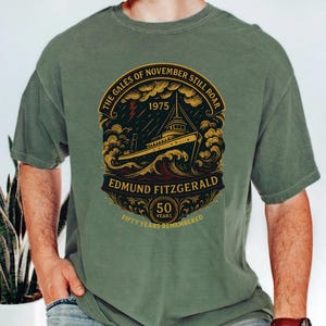 May include: Olive green t-shirt featuring a detailed graphic of a ship at sea, with the text "The Gales of November Still Roar" and "Edmund Fitzgerald". The design includes the year 1975 and "50 Years Remembered".
