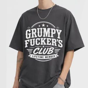May include: Dark grey t-shirt with the text "GRUMPY FUCKER'S CLUB LIFETIME MEMBER" in white. The graphic is centered on the front of the shirt. The shirt has short sleeves and a crew neck.
