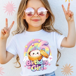 Pu&ograve; includere: Una t-shirt bianca a maniche corte con una grafica colorata che raffigura un raviolo in un cestello di bamb&ugrave;, un arcobaleno e il numero 6. La scritta Birthday Girl &egrave; in un carattere giocoso, circondata da stelle, nuvole e cuori pastello.