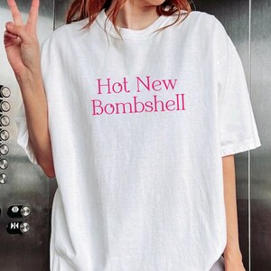 May include: A white short-sleeved t-shirt featuring the text "Hot New Bombshell" printed in a bold, dark pink font across the center of the chest. The garment has a crew neckline and a relaxed, loose-fitting silhouette with standard hemmed sleeves.