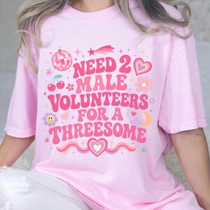 May include: A light pink t-shirt featuring a graphic print with the text "NEED 2 MALE VOLUNTEERS FOR A THREESOME" in a bold, wavy pink font. The design includes colorful illustrations of a disco ball, cherries, a heart, a flower, and a star.