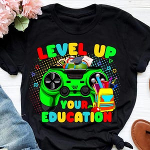 Könnte beinhalten: Schwarzes T-Shirt mit farbenfroher Grafik. Das Design zeigt einen grünen Videospiel-Controller mit den Worten "LEVEL UP YOUR EDUCATION" in hellen, fetten Buchstaben. Ein Rucksack und eine Abschlusskappe sind ebenfalls enthalten.