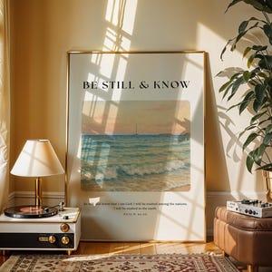May include: A framed art print with the text "BE STILL & KNOW" above a seascape painting. The print is in a gold frame, and the scene includes a sailboat on the water. A vintage record player and a potted plant are also in the room.
