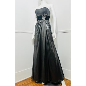Puede incluir: Un vestido de gala plateado metálico sin tirantes con una banda de cintura de satén negro y un adorno de pedrería plateada. El vestido tiene una falda fluida y un corpiño ajustado.
