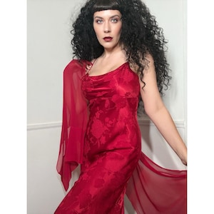 May include: A red, floral-patterned slip dress with spaghetti straps and a cowl neckline. A matching sheer red shawl is draped over the shoulders. The dress is floor-length and has a side slit.