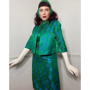 May include: A vintage green floral print dress, jacket, and headscarf set. The set features a vibrant green fabric with blue floral patterns. The jacket has three-quarter sleeves, and the dress is knee-length. The ensemble is completed with long white gloves.