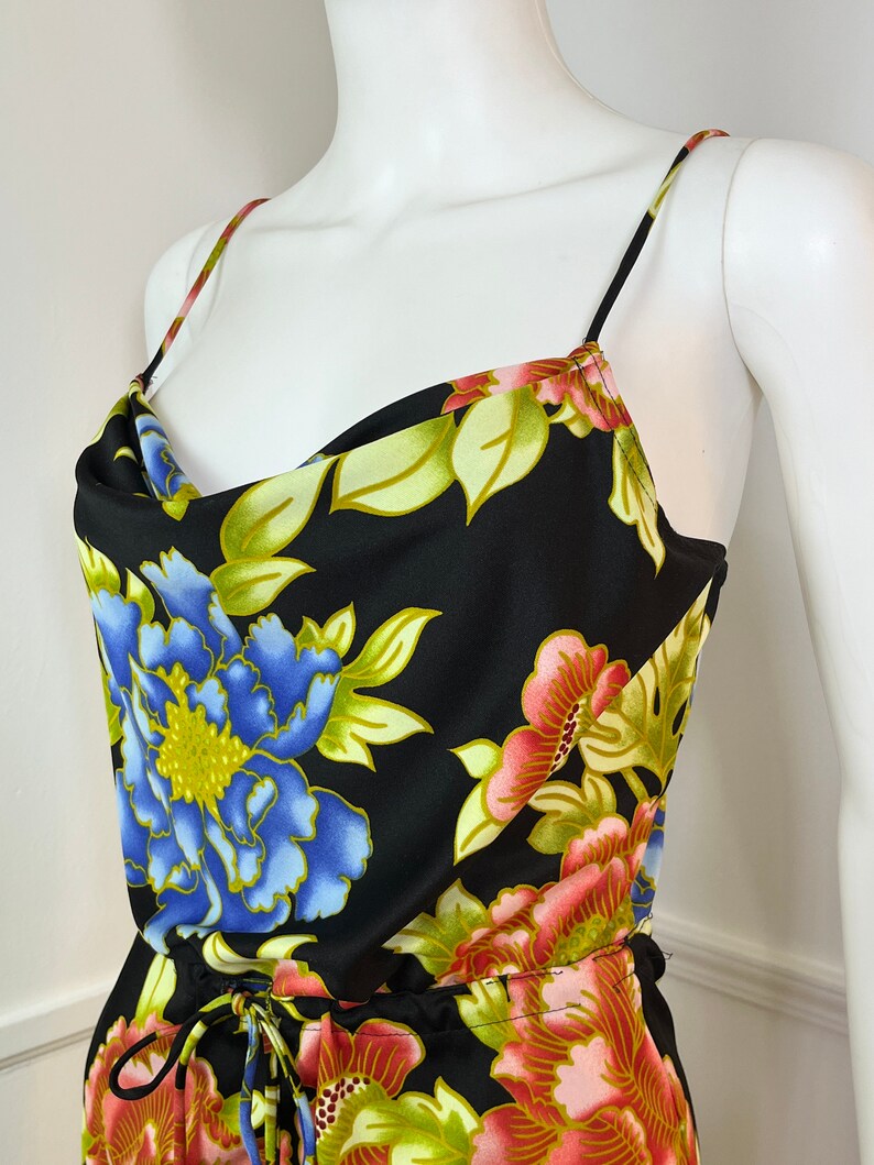 May include: A black floral print dress with a cowl neck and thin straps. The dress has a drawstring waist and a large floral print in shades of blue, pink, green, and yellow.