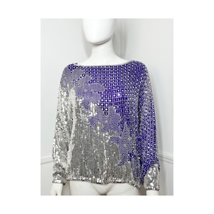 Puede incluir: Una blusa de lentejuelas de color morado y plateado con un diseño floral. La blusa tiene mangas largas y un escote redondo.